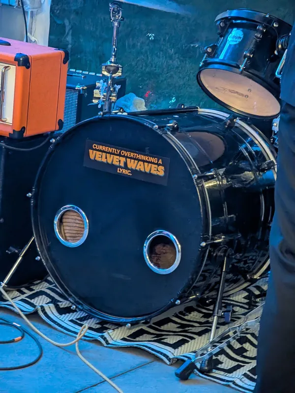 The band's kick drum seen from floor level, its black head carrying an orange and white sticker reading CURRENTLY OVERTHINKING A VELVET WAVES LYRIC. An orange amplifier head sits at the left, a floor tom and cymbal stand at the right, and the kit rests on a black and white patterned rug under blue stage light.