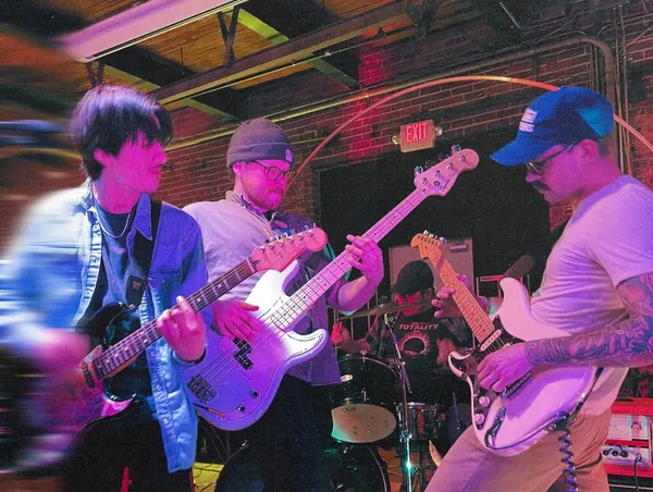 Three Velvet Waves players crowded together at the front of a brick-walled room under magenta and purple light, guitar necks crossing and the whole frame smeared with motion blur, with the drummer visible between them at the back.