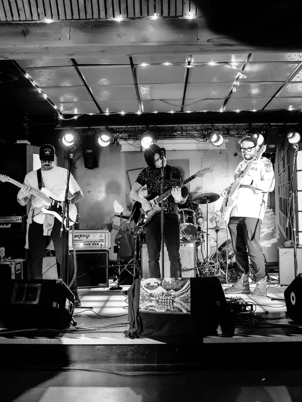 Black and white wide shot of Velvet Waves on the King's Rook Club stage under a ceiling strung with lights: bassist at left, singer and guitarist at the centre mic, second guitarist at right, and the drummer seated behind them. A banner carrying the band's name is draped across the front of the stage.