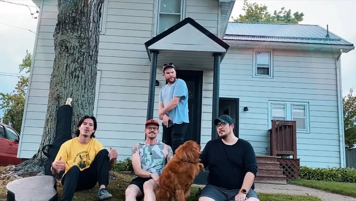 The four members of Velvet Waves outside a pale blue clapboard house on a summer evening: three sitting on the grass under a big tree with a golden retriever between them, the fourth standing on the porch step behind.