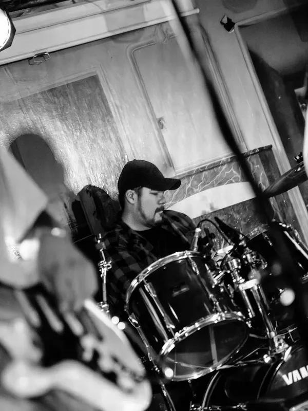 Black and white shot of Sean behind the drum kit at King's Rook Club, in a baseball cap and plaid shirt, head down and mid-stroke, with the bass drum and cymbal stands filling the foreground.