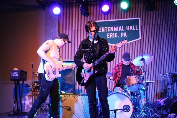 Velvet Waves playing beneath a banner reading CENTENNIAL HALL ERIE, PA: the bassist leaning back at left, the singer at the centre microphone with a guitar, and the drummer in a red plaid shirt at the kit on the right, under teal and purple spotlights.
